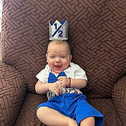 Christian joined the competition — help win amazing prizes! baby, child, smiling, crown, half_birthday, blue_shorts, white_shirt, bow_tie, armchair, brown_furniture, happy, celebration, indoor, cute, person, infant, sitting, portrait, playful, festive