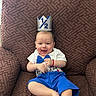 baby, child, smiling, crown, half_birthday, blue_shorts, white_shirt, bow_tie, armchair, brown_furniture, happy, celebration, indoor, cute, person, infant, sitting, portrait, playful, festive