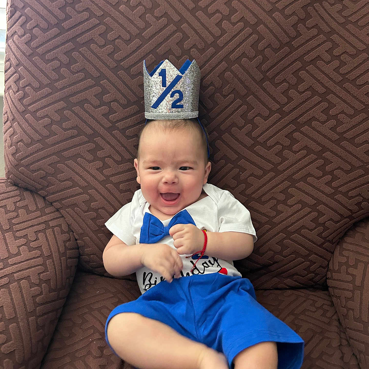 Christian joined the competition — help win amazing prizes! baby, child, smiling, crown, half_birthday, blue_shorts, white_shirt, bow_tie, armchair, brown_furniture, happy, celebration, indoor, cute, person, infant, sitting, portrait, playful, festive