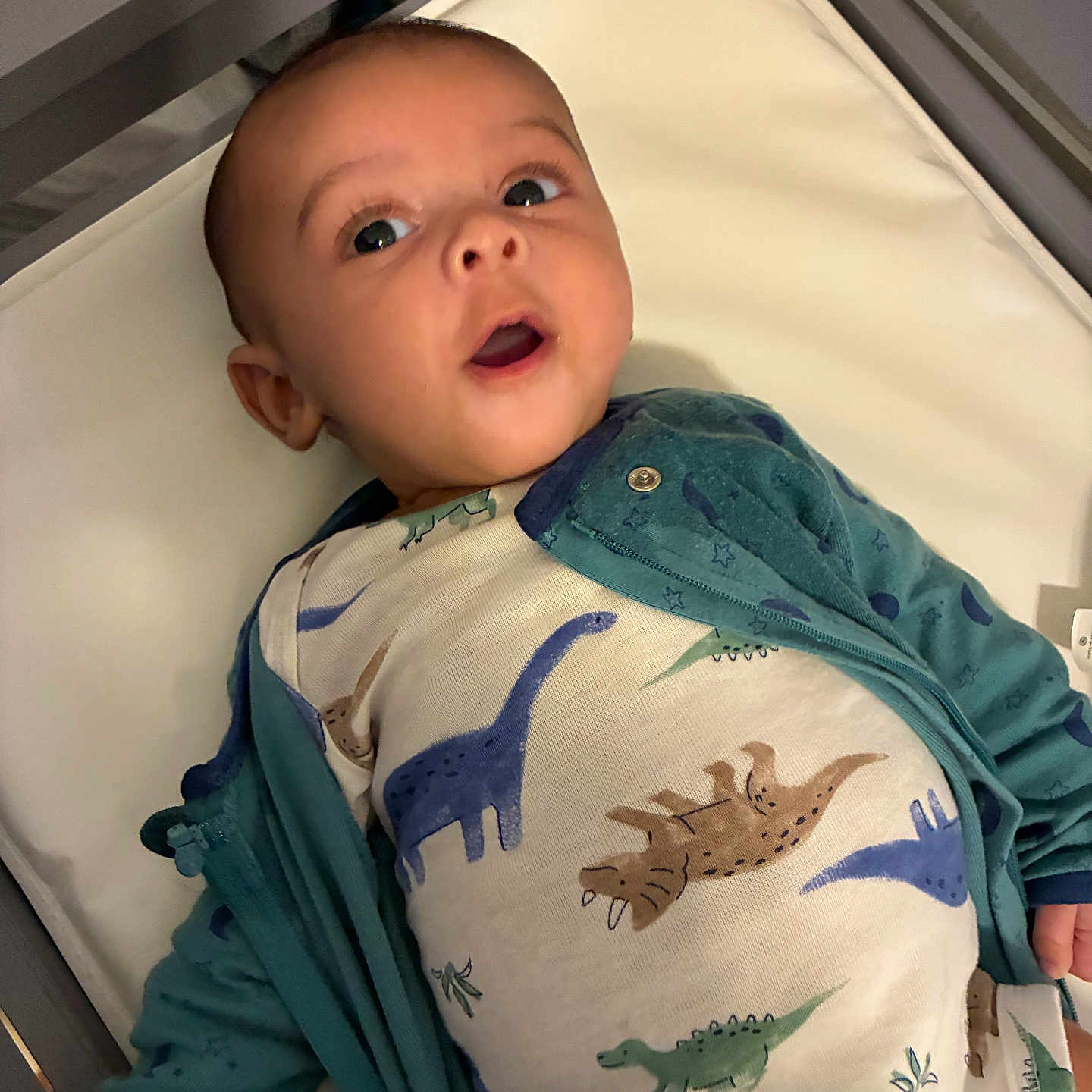 Orion is registered to the contest to win money with this photo: adorable, baby, bed, child, clothing, crib, cute, dinosaur_print, expression, face, indoor, infant, jacket, lying_down, mouth_open, onesie, pale_skin, small_hand, soft_surface, wide_eyes