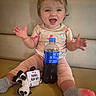 toddler, child, smiling, dinosaur_shirt, socks, plush_toy, cow_toy, soda_bottle, bench, cushion, happy, indoor, baby, hands_up, casual_clothing, dried_flowers, cute, person, portrait, playful