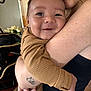 baby, child, smiling, hug, person, adult, tattoo, brown_clothing, indoor, home, window_blinds, flower_vase, hand, face, portrait, close_up, happy, skin, arm, candid