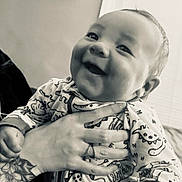 Kailo is registered to the contest to win money with this photo: baby, child, smiling, tattoo, hand, onesie, dinosaur_pattern, indoor, person, happy, infant, clothing, portrait, face, cute, holding, home, black_and_white, cozy, closeup
