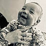 baby, child, smiling, tattoo, hand, onesie, dinosaur_pattern, indoor, person, happy, infant, clothing, portrait, face, cute, holding, home, black_and_white, cozy, closeup