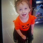 Amora joined the competition — help win amazing prizes! child, girl, smiling, orange_dress, halloween, pumpkin_face, playful, indoors, floor, tiles, motor_oil, container, shelf, cute, person, young_child, hair_tied, footwear, tongue_out, happy