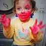 toddler, child, paint, red_paint, face_paint, hands, yellow_sweater, bird_design, blue_pants, sink, mirror, indoor, messy, playful, curly_hair, smiling, expression, person, home, cabinet