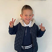 Mario participe au concours pour gagner de l'argent avec cette photo : belt, boy, casual_clothing, child, cute, fairy_lights, fashion, gesture, happy, hoodie, indoor, peace_sign, person, playful, portrait, short_hair, smile, standing, vest, white_wall