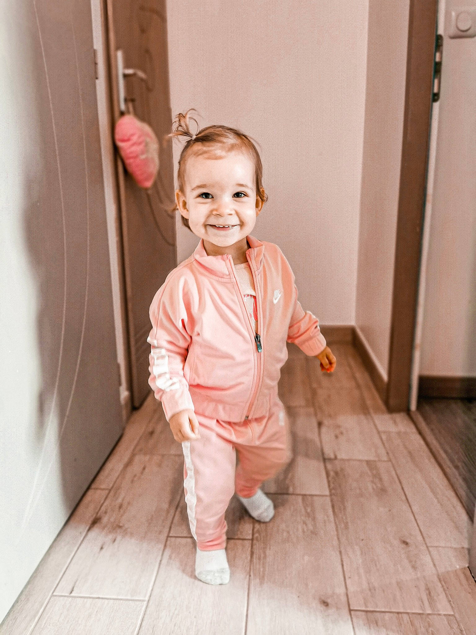 Malia a rejoint le concours — aidez-le/la à gagner de superbes lots ! baby, baby_toddler_clothing, cheek, door, eye, floor, flooring, happy, hardwood, human_body, joy, nose, person, pink, sleeve, smile, standing, toddler, varnish, wood