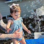 Antoinette joined the competition — help win amazing prizes! child, toddler, bubbles, tattoo, hand, blue_clothing, bow, outdoor, sunlight, playtime, baby, person, skin, summer, joy, fun, background, house, blur, happy