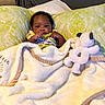 baby, blanket, pillow, plush_toy, child, indoor, portrait, cute, infant, cozy, bed, smiling, furniture, person, soft, patterned, clothing, relaxing, young_child, comfort