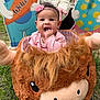 baby, child, pink_headband, pink_clothing, plush, stuffed_animal, grass, outdoor, hand, person, cute, smiling, face, head, toy, playful, welcome_sign, colorful, sitting, young_child