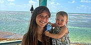 Everlyn joined the competition — help win amazing prizes! woman, toddler, child, smile, outdoor, ocean, beach, sunny, gazebo, wooden_structure, water, sky, rocks, holding, happy, casual_clothing, nature, vacation, portrait, family