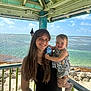 woman, toddler, child, smile, outdoor, ocean, beach, sunny, gazebo, wooden_structure, water, sky, rocks, holding, happy, casual_clothing, nature, vacation, portrait, family