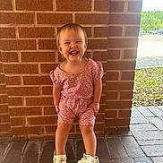 Everlyn is registered to the contest to win money with this photo: toddler, child, smiling, standing, pink_clothing, romper, crocs, unicorn, footwear, brick_wall, outdoor, sunny, happy, person, cute, short_hair, greenery, pavement, daylight, parking_lot