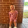 toddler, child, smiling, standing, pink_clothing, romper, crocs, unicorn, footwear, brick_wall, outdoor, sunny, happy, person, cute, short_hair, greenery, pavement, daylight, parking_lot