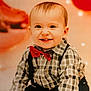 baby, child, smiling, tooth, red_bow_tie, checkered_shirt, suspenders, sitting, fluffy_rug, indoor, portrait, cute, happy, face, hands, feet, balloons, lights, warm_colors, celebration
