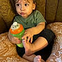 toddler, child, baby, sippy_cup, cushion, pattern, green, orange, face, expression, curious, sitting, clothing, short_sleeve, indoor, foot, hand, brown, hair, comfort