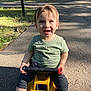 toddler, child, smiling, outdoor, toy_vehicle, green_grass, sunlight, trees, park, happy, casual_clothing, blue_shoes, short_sleeve_shirt, pavement, daytime, nature, play, cute, young_child, fun