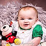 baby, infant, child, portrait, smiling, plush_toy, mickey_mouse, stuffed_animal, fur_blanket, studio_photography, closeup, dress, lace, green_clothing, basket, eyes, rosy_cheeks, soft_texture, watermark, happy
