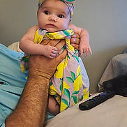 Emma is registered to the contest to win money with this photo: arm, baby, blanket, child, colorful, cozy, curious, dress, expression, furniture, hand, headband, holding, indoor, lemon, pattern, person, remote_control, skin, wide_eyes