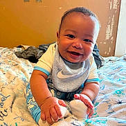 Jaevon is registered to the contest to win money with this photo: baby, bed, bedroom, blanket, child, close_up, clothing, feet, hands, happy, home, indoor, infant, nike_shirt, patterned_blanket, portrait, sitting, smiling, socks, warm_lighting