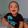 baby, bed, black_shirt, blanket, child, clothing, cozy, cute, dinosaur, face, hand, happy, indoors, infant, pillow, portrait, room, sitting, smiling, young