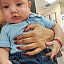 baby, child, infant, person, hand, clothing, denim, shorts, blue_shirt, nail_polish, red_nails, bracelet, indoor, office, wall, documents, supporting_hand, seated, curious, calm