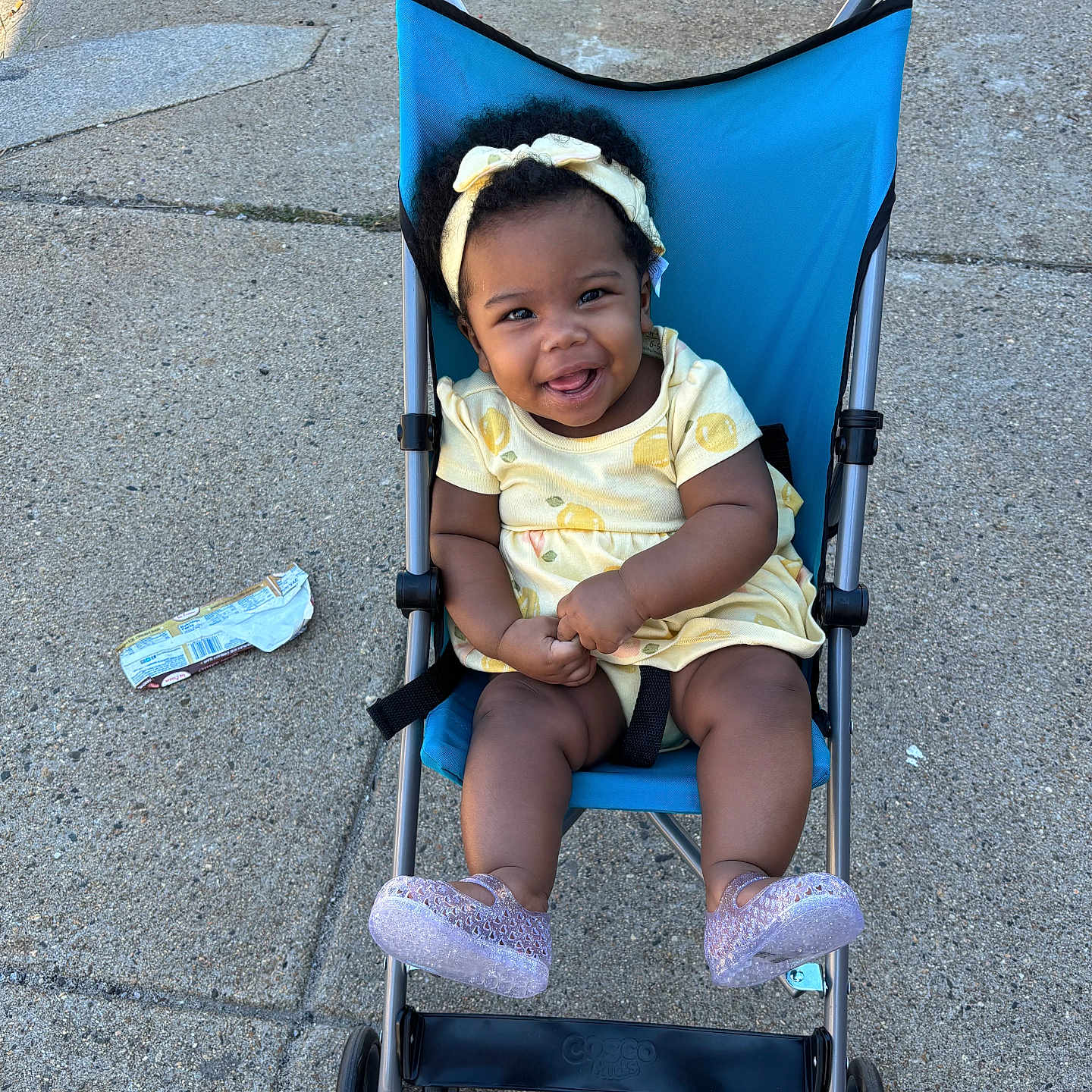 Naela joined the competition — help win amazing prizes! baby, child, stroller, smiling, outdoor, pavement, sidewalk, bow, headband, yellow_outfit, dress, shoes, seated, infant, portrait, happy, sunlight, urban, parked_stroller, gummy_smile
