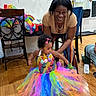 child, toddler, infant, adult, woman, smiling, laughing, rainbow_dress, colorful_tutu, pink_bow, birthday, indoor, family, celebration, wooden_floor, chair, balloons, party, portrait, home