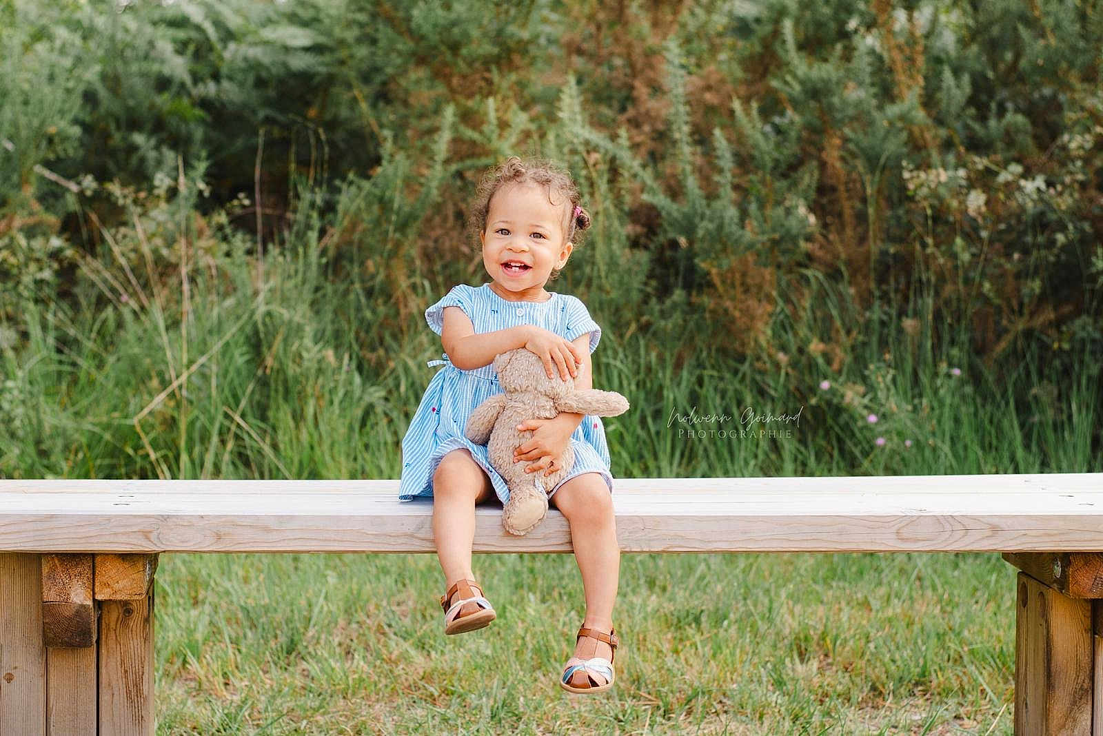 Zohra participe au concours pour gagner de l'argent avec cette photo : toddler, child, smiling, blue_dress, teddy_bear, bench, outdoor, greenery, grass, happy, cute, sandals, person, nature, daylight, portrait, young_child, sitting, toy, playful