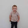 child, boy, standing, beige_shirt, black_pants, boots, wall, concrete, outdoor, casual_clothing, portrait, curious_expression, short_hair, young, person, footwear, standing_pose, neutral_background, casual_style, daylight