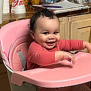 Arya joined the competition — help win amazing prizes! baby, high_chair, pink, smiling, indoor, child, curly_hair, kitchen, snacks, stacked_items, wooden_counter, tableware, person, cute, happy, sitting, clothing, long_sleeves, polka_dots, infant