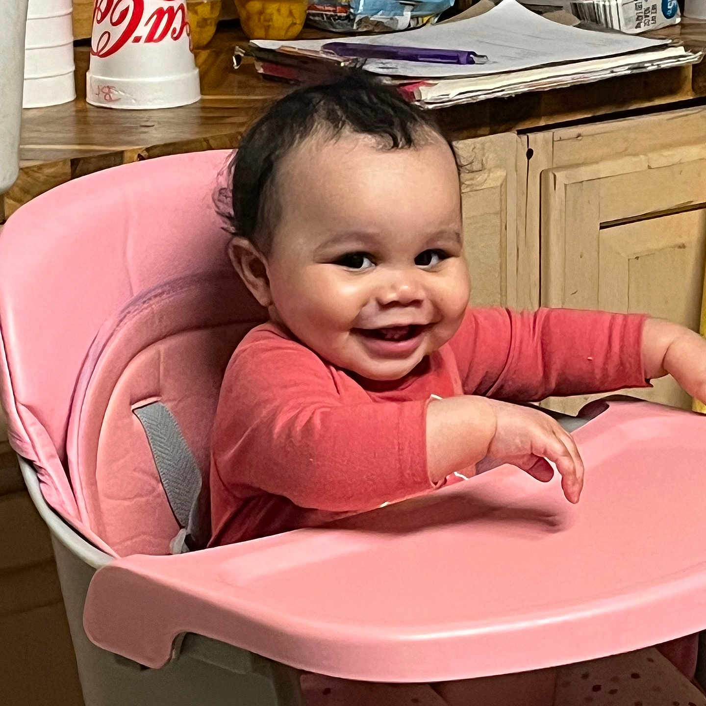 Arya joined the competition — help win amazing prizes! baby, high_chair, pink, smiling, indoor, child, curly_hair, kitchen, snacks, stacked_items, wooden_counter, tableware, person, cute, happy, sitting, clothing, long_sleeves, polka_dots, infant