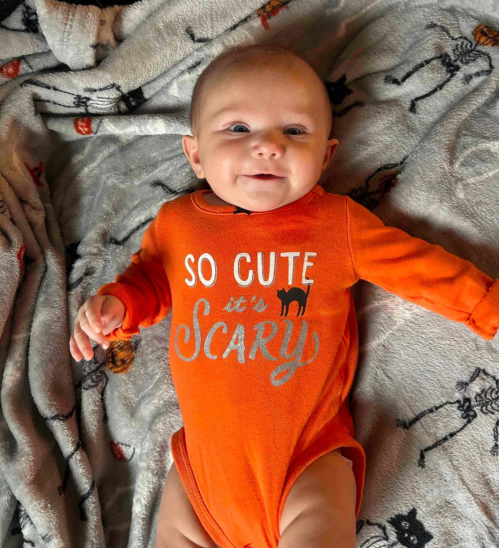 Julie is registered to the contest to win money with this photo: baby, infant, orange_onesie, smiling, blanket, skeleton_pattern, black_cat_pattern, cute, lying_down, cozy, indoors, child, face, hand, head, expression, holiday_theme, halloween, soft_texture, person