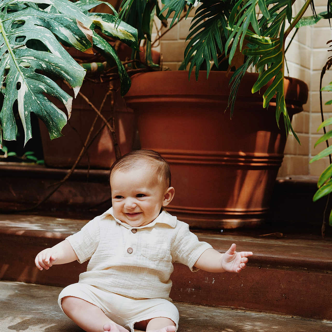 Lorenzo joined the competition — help win amazing prizes! baby, barefoot, casual_clothing, child, concrete_floor, cute, greenery, happy, indoors, infant, natural_light, plants, playful, portrait, shirt, shorts, sitting, smiling, terracotta_pot, toddler