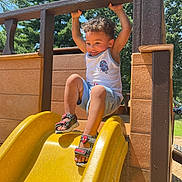 Damian is registered to the contest to win money with this photo: toddler, child, playground, slide, yellow_slide, outdoor, summer, smiling, happy, sandals, shorts, tank_top, curly_hair, trees, wooden_structure, sunlight, play, fun, park, nature