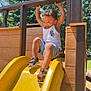toddler, child, playground, slide, yellow_slide, outdoor, summer, smiling, happy, sandals, shorts, tank_top, curly_hair, trees, wooden_structure, sunlight, play, fun, park, nature