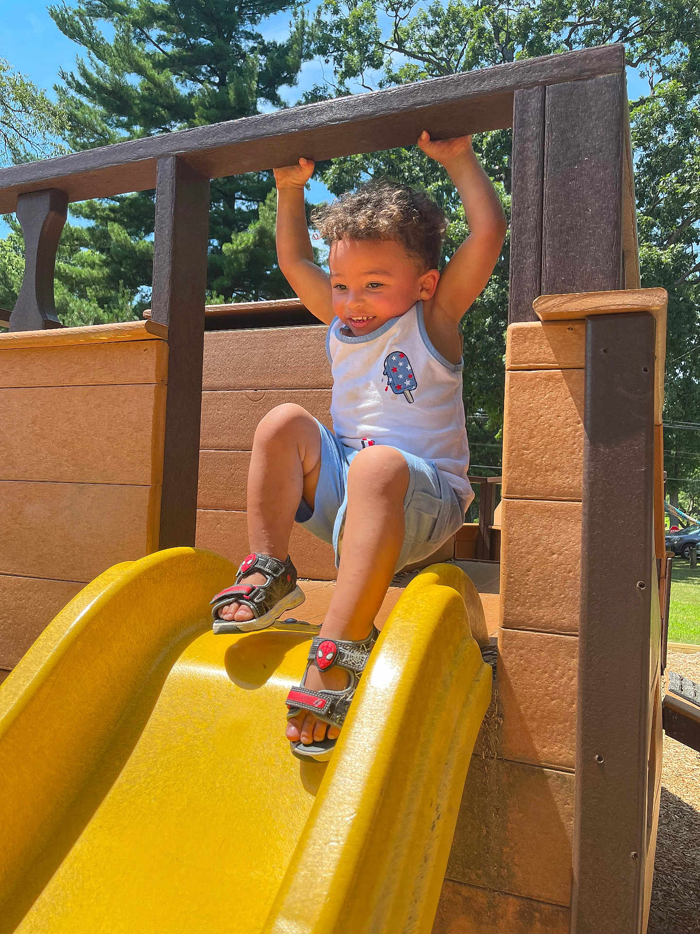 Damian is registered to the contest to win money with this photo: toddler, child, playground, slide, yellow_slide, outdoor, summer, smiling, happy, sandals, shorts, tank_top, curly_hair, trees, wooden_structure, sunlight, play, fun, park, nature