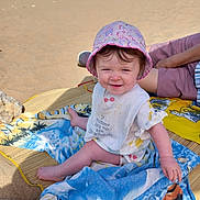 Elizabeth joined the competition — help win amazing prizes! baby, child, beach, sand, towel, hat, pink_hat, bib, smile, sunny, sea, water, person, outdoor, summer, relaxation, vacation, clothing, happy, family