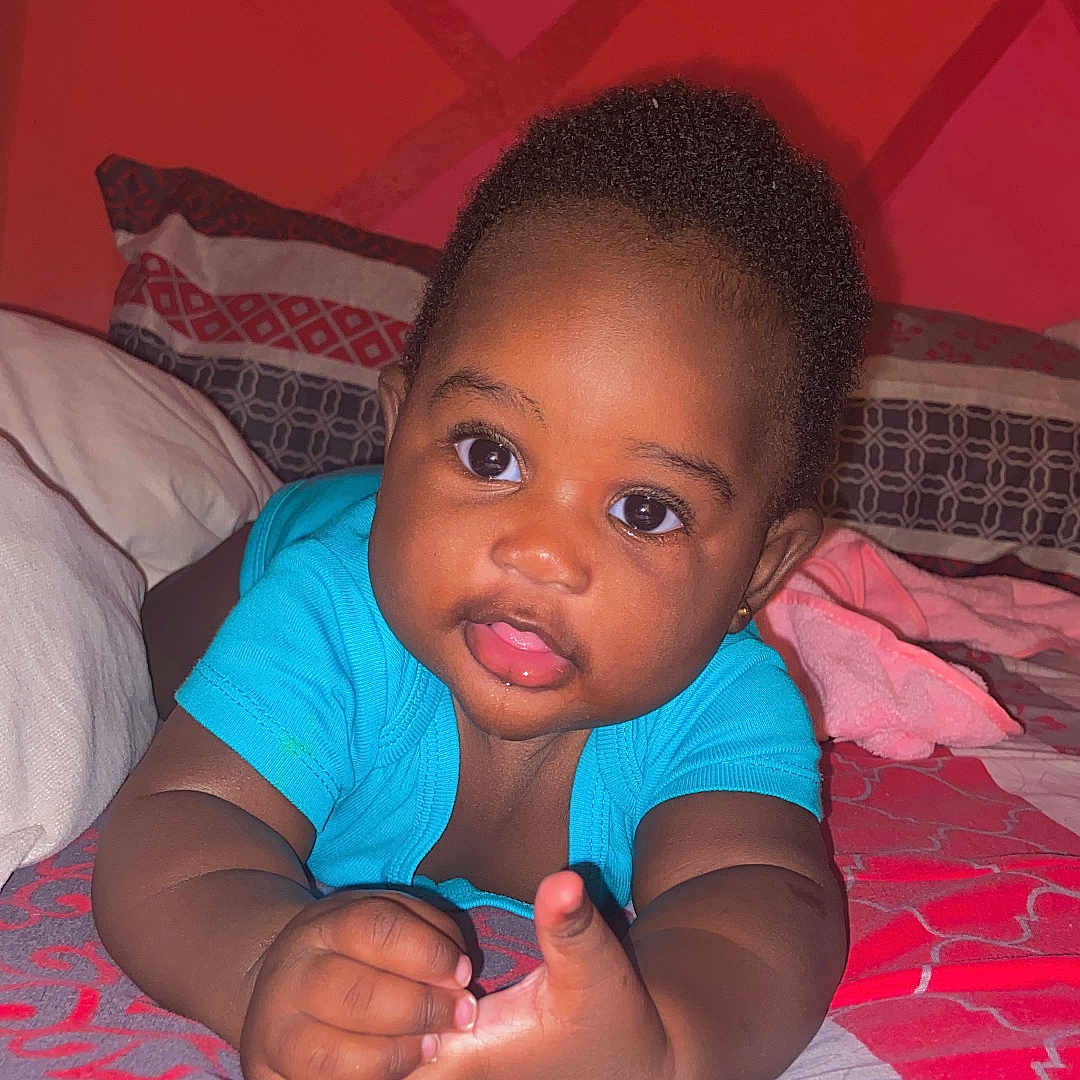 Kiana Lewis is registered to the contest to win money with this photo: baby, child, infant, blue_clothing, bed, pillow, red_wall, geometric_pattern, curious_expression, lying_down, indoors, portrait, cute, smiling, wide_eyes, home, colorful, patterned_bedspread, young_child, cozy