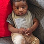 Braxton is registered to the contest to win money with this photo: baby, child, infant, couch, pillow, gray_clothing, bib, beige_pants, bear_print, curly_hair, indoor, seated, cute, expression, hands, tongue_out, soft_lighting, home, comfortable, portrait