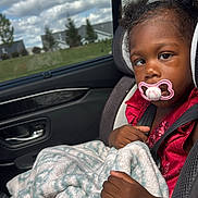Amari joined the competition — help win amazing prizes! child, toddler, car_seat, pacifier, blanket, curly_hair, red_dress, car_interior, window, clouds, sky, trees, suburban, seatbelt, hand, face, portrait, daytime, expression, safety