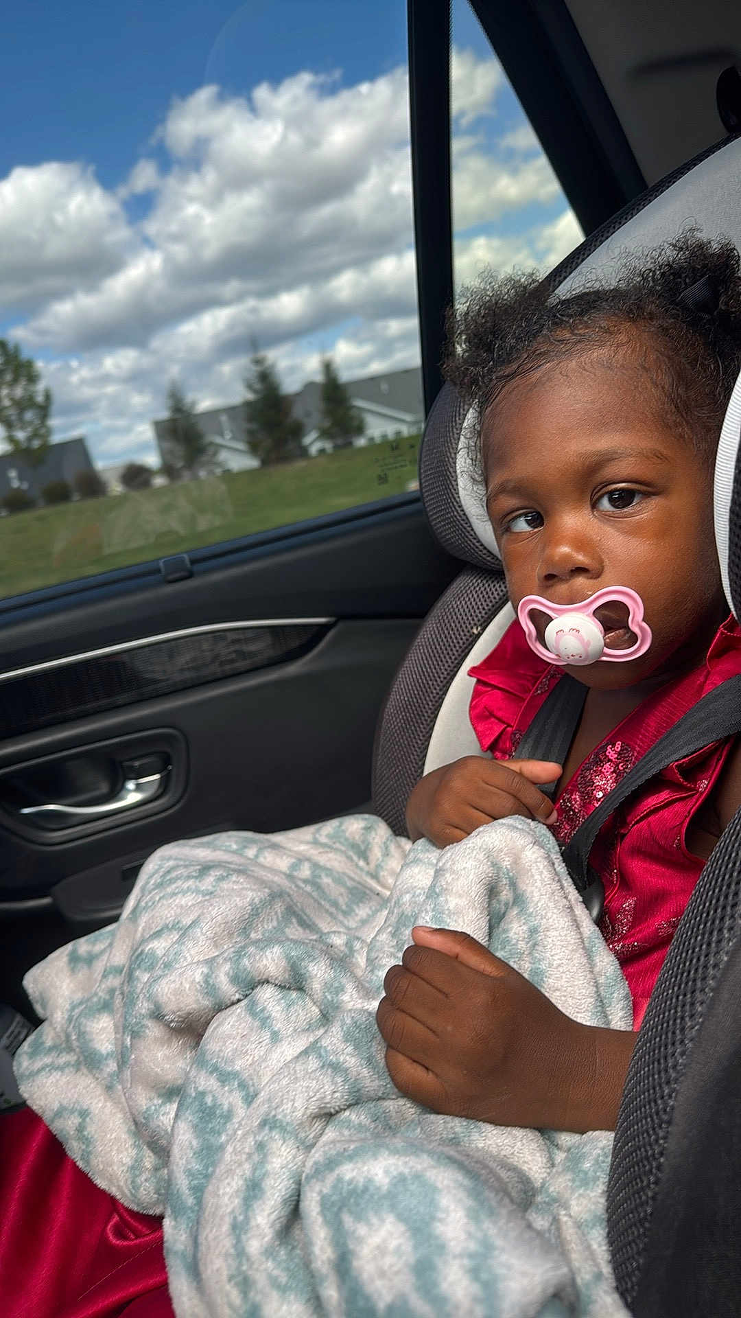 Amari joined the competition — help win amazing prizes! child, toddler, car_seat, pacifier, blanket, curly_hair, red_dress, car_interior, window, clouds, sky, trees, suburban, seatbelt, hand, face, portrait, daytime, expression, safety