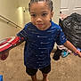 barefoot, black_shorts, blue_shirt, boy, braided_hair, carpet, champion_brand, child, clothes, door, home, indoor, person, plastic_bag, playful, stairs, standing, toy_shield, wall, watch