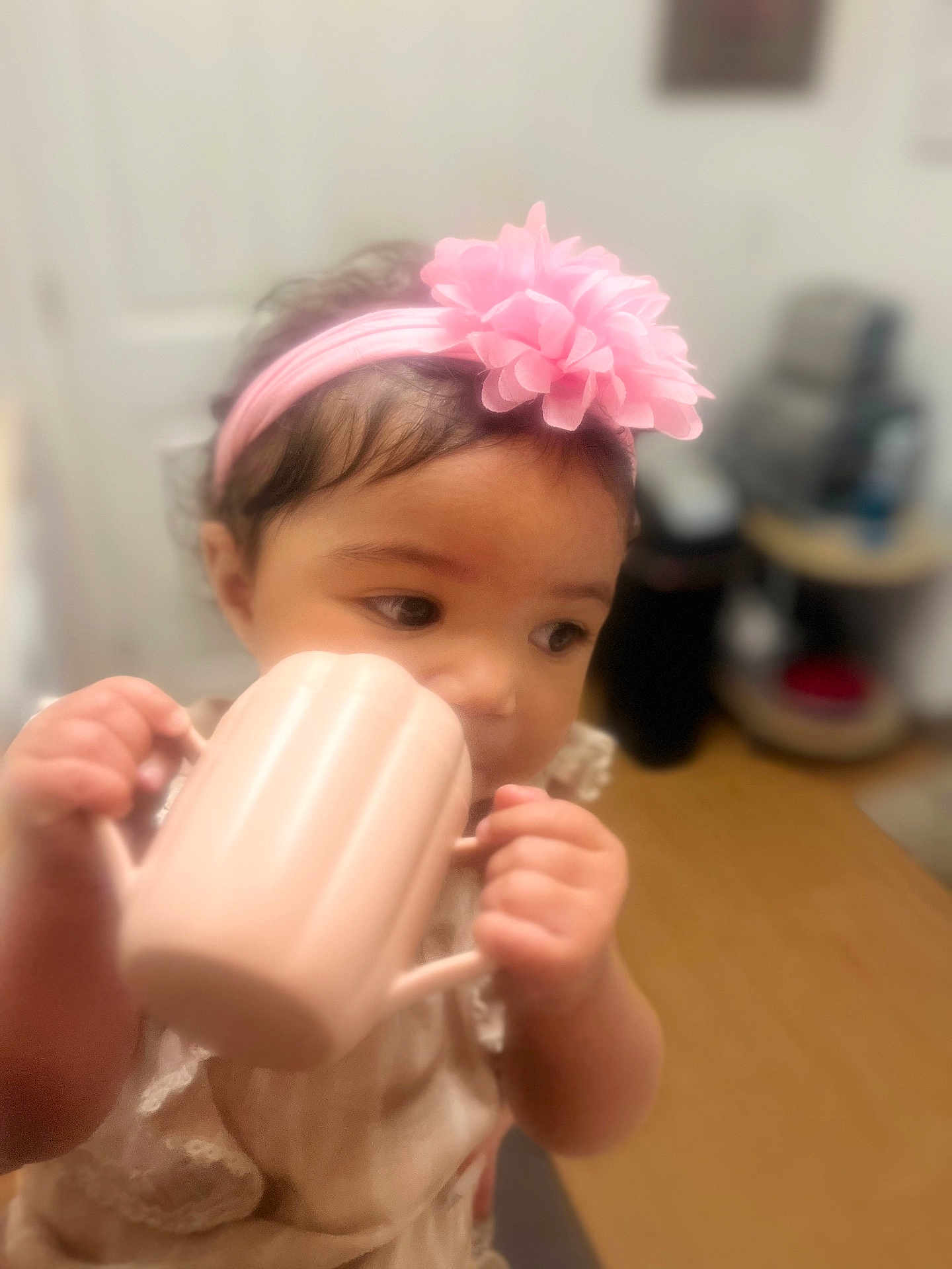 Braelynn is registered to the contest to win money with this photo: toddler, child, pink_headband, flower, mug, hands, indoor, table, curious, face, clothing, person, portrait, soft_focus, baby, headwear, drinking, cute, young_child, furniture