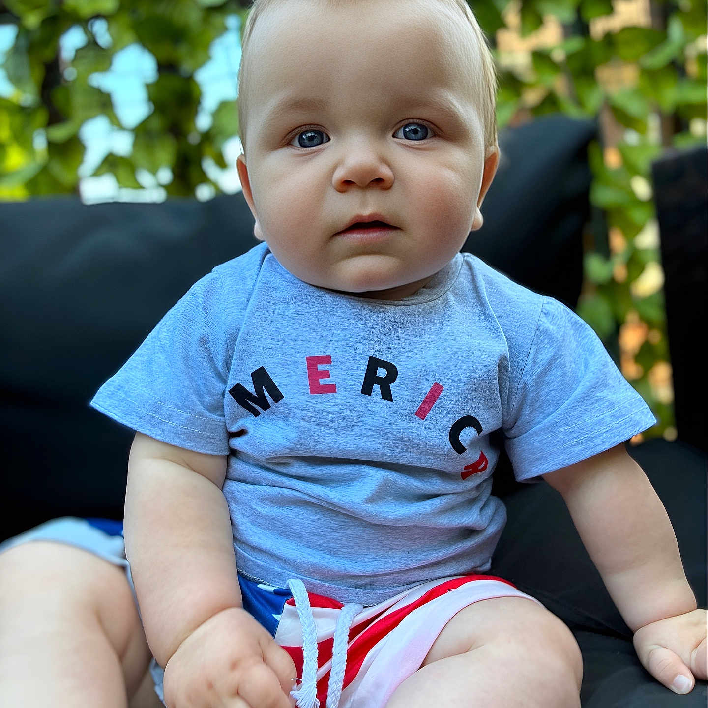 Kruz joined the competition — help win amazing prizes! baby, black_cushion, blue_eyes, casual_clothing, child, cushion, cute, expression, face, greenery, hand, infant, legs, outdoor, portrait, shorts, sitting, summer, tshirt, young