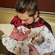 Brycen is registered to the contest to win money with this photo: newborn, infant, baby, adult, holding, swaddled, hospital_blanket, blanket, wristband, hospital_room, bench, cuddle, portrait, close_up, hair_bun, facial_hair, red_black_shirt, skin, parenthood, love