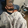 baby, newborn, man, beard, cap, blanket, hospital_room, recliner, medical_cart, sleeping, holding, bonding, swaddle, cozy, portrait, chair, wood_floor, equipment, cap_logo, patient_room
