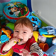 Jules a rejoint le concours — aidez-le/la à gagner de superbes lots ! toddler, child, toy_car, red_shirt, striped_sleeves, indoor, play_area, colorful_toys, mickey_mouse_balloon, happy, curious, reaching_hand, baby, playtime, smiling, face, hair, toy, plastic_toys, floor_mat