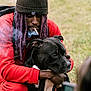 person, dog, smoking, beanie, dreadlocks, outdoor, grass, black_dog, red_clothing, pet, animal, smoke, crouching, leash, closeup, portrait, casual, human_animal_interaction, nature, calm